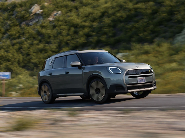Side view of a driving all-electric MINI Countryman in Smokey Green with Vibrant Silver Roof off-road.