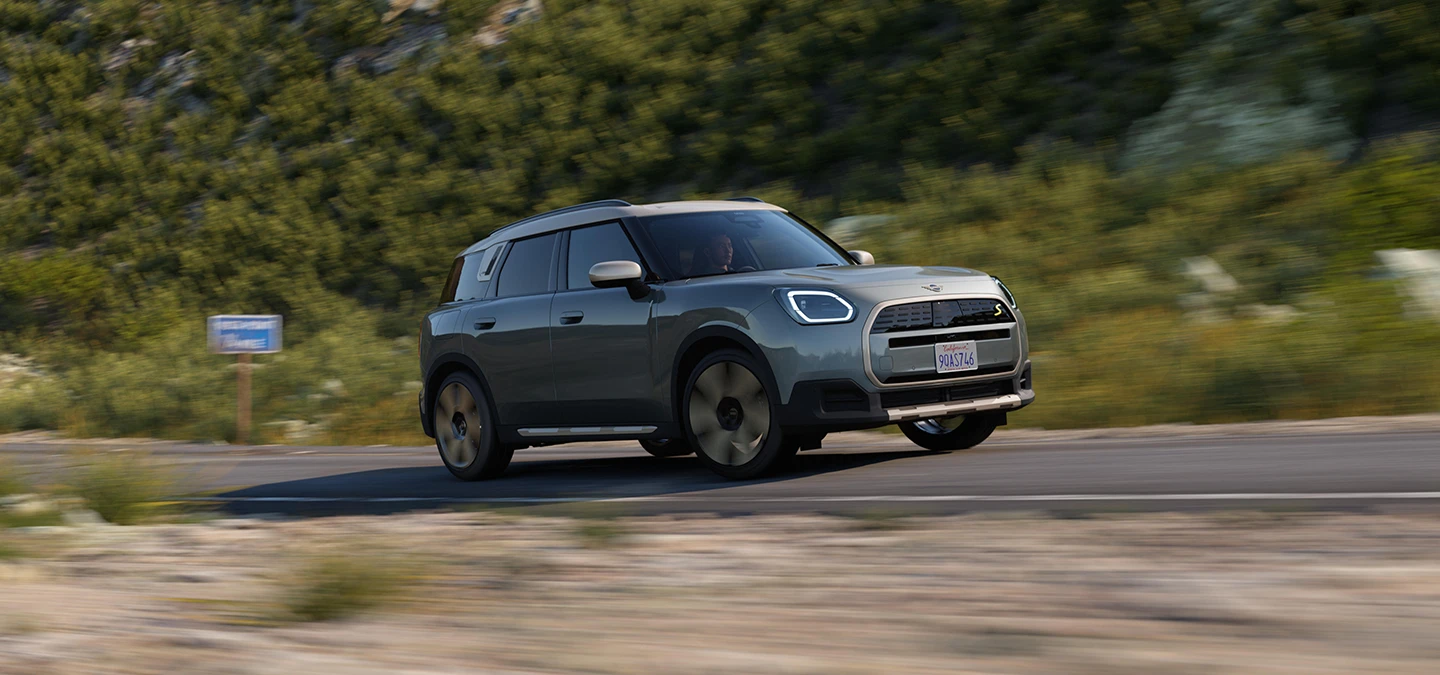 Side view of a driving all-electric MINI Countryman in Smokey Green with Vibrant Silver Roof off-road.