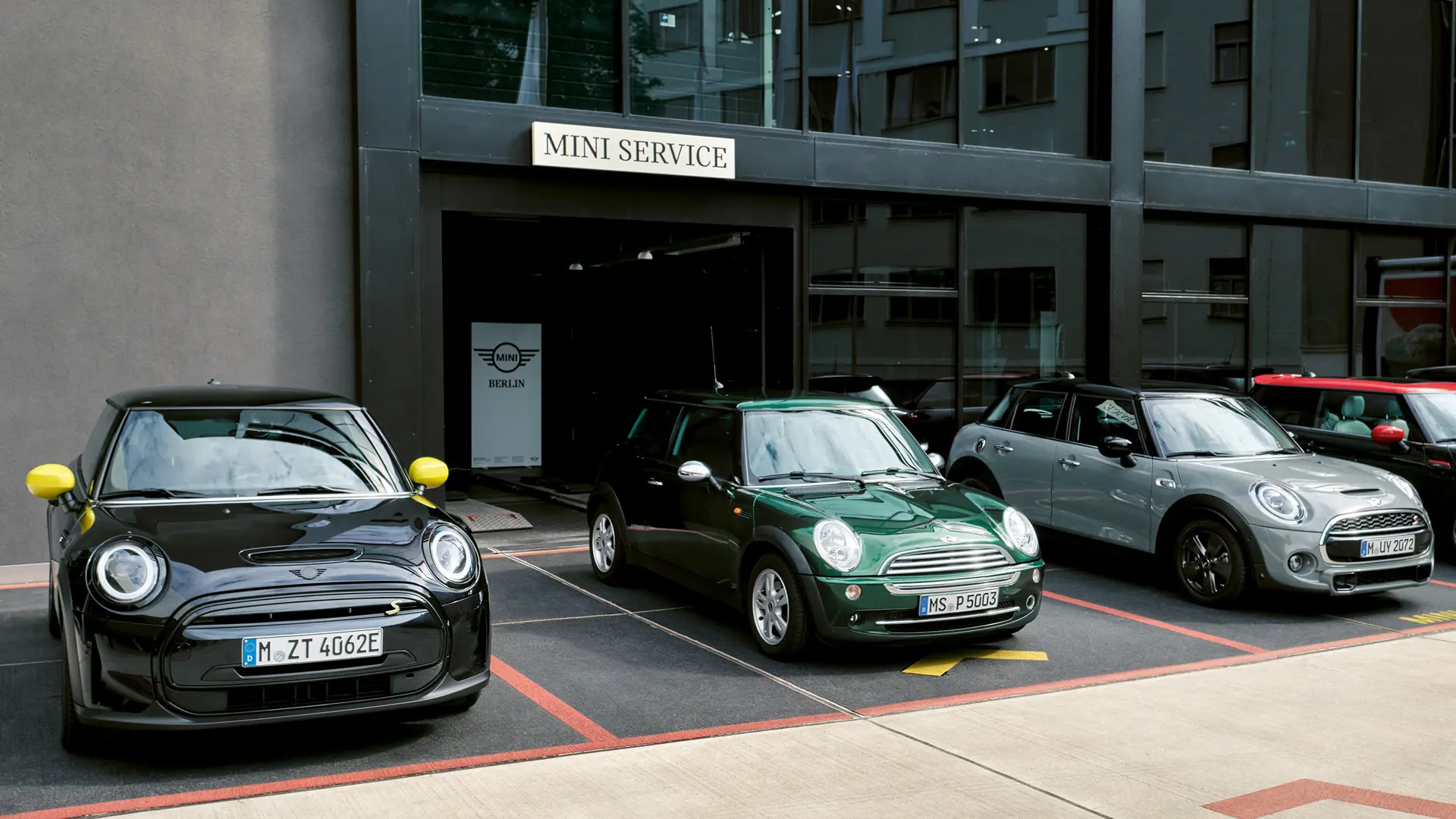 MINI Cooper Electric S in black standing outside a MINI Centre alongside a MINI Cooper 3-door in green, MINI 5-door in silver, MINI JCW black, front view. 